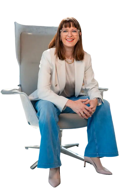 Attraktive Frau sitzt auf einem modernen Stuhl, lächelt freundlich und wirkt selbstbewusst. Sie trägt eine weiße Bluse, einen hellen Blazer und blaue Jeans, was Professionalität und Zugänglichkeit ver.
